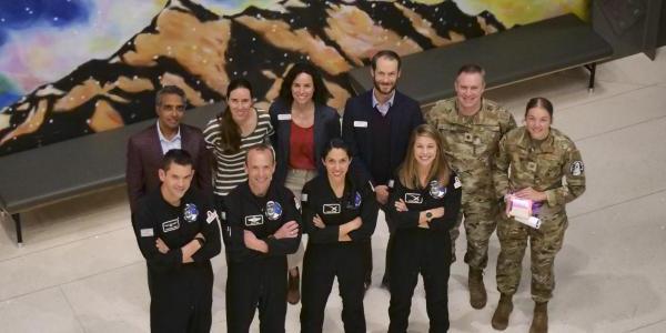 10 people, four wearing black flight suits and tow wearing camouflage uniforms, pose for a photo as seen from above